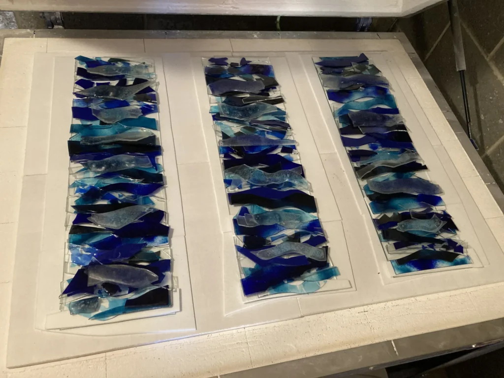 Three rectangular glass panels, each filled with layered pieces of blue, teal, and clear fused glass in wave-like or organic shapes. The panels are laid flat on a white surface, likely inside a kiln, ready for the fusing process.