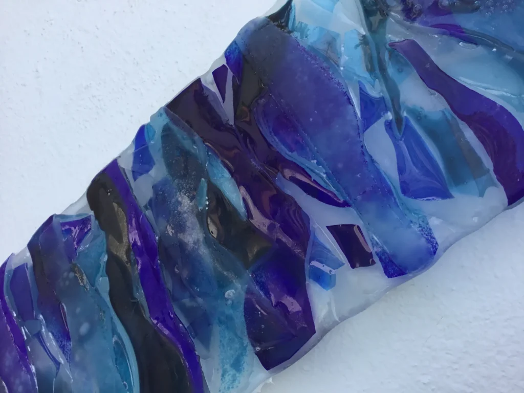 A close-up view of a fused glass art panel, showing layered pieces of deep blue, lighter blue, teal, and some dark grey or black glass. The pieces are irregularly shaped, creating a fluid, wave-like pattern across the surface, with some areas appearing slightly textured or bubbled from the fusing process