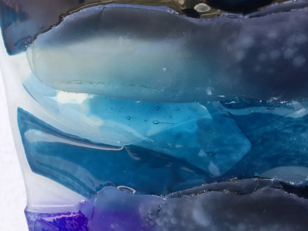 An extreme close-up view of a fused glass art panel, highlighting horizontal layers of clear, light blue, and deep blue glass. The glass has an organic, wavy form, with visible texture and small trapped air bubbles, enhancing the water-like appearance.