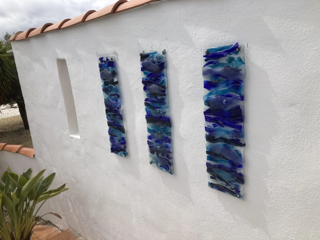 Three rectangular, textured fused glass art panels, predominantly in shades of blue and teal, mounted vertically on a white stucco exterior wall. The glass pieces are layered to create a wavy, water-like appearance, catching the light. The wall has a terracotta tiled top edge, and a plant is visible in the foreground.