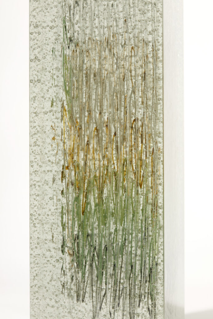 A vertical close-up of the side of the "Memories of Margate" glass artwork. The clear glass block is heavily textured with numerous small bubbles. Through the side, the internal, vertical strands of glass are visible, appearing as thin, wavy lines. The colors of these strands transition from a lighter, almost brownish-yellow at the top to various shades of green towards the bottom