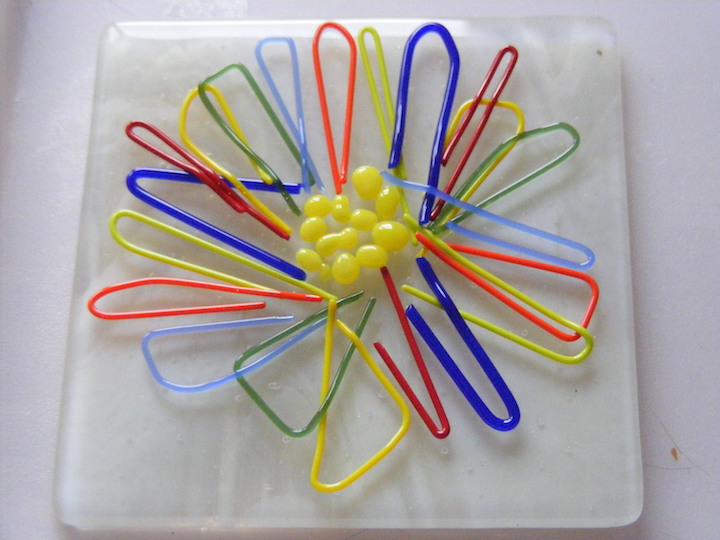 A close-up of a single daisy made from fused red, blue, green, and yellow glass loops.