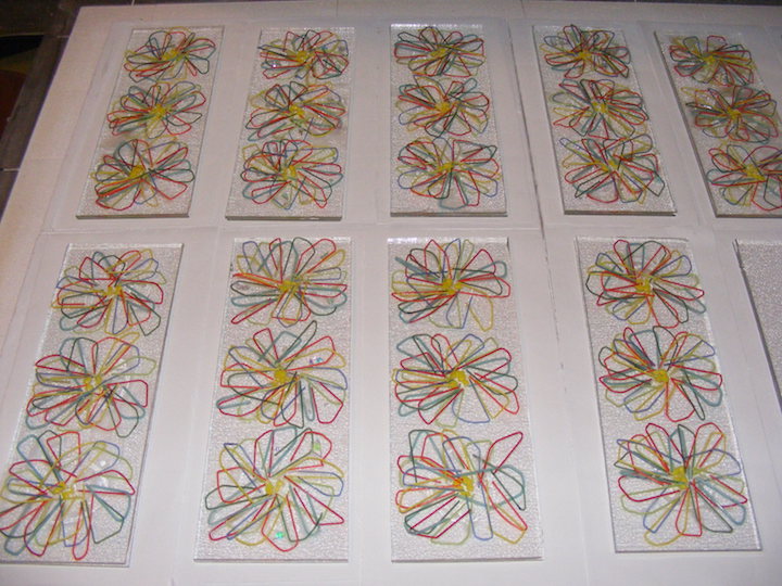 A grid of flat glass panels, each with a colourful looped daisy pattern, laid out during the assembly process.