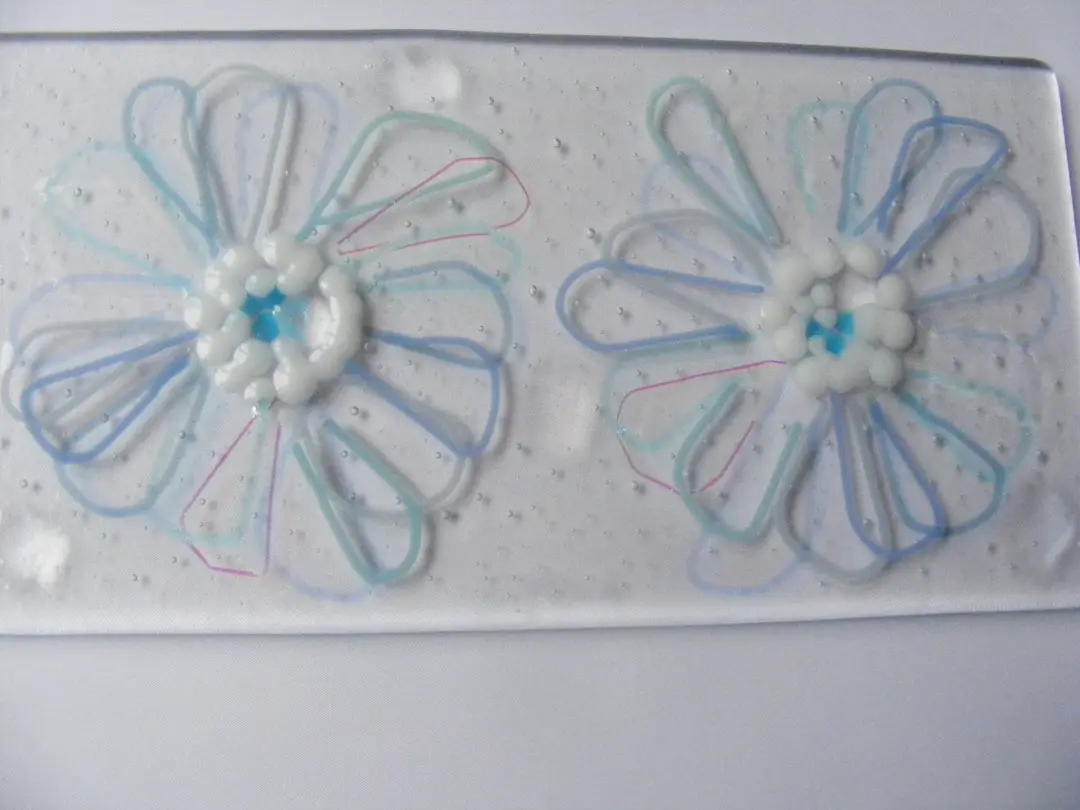 A flat, light blue glass panel with two white and clear layered daisy patterns fused onto its surface.
