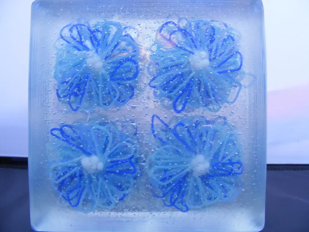 A close-up of a glass block, showcasing four intricate blue daisies with visible air bubbles.