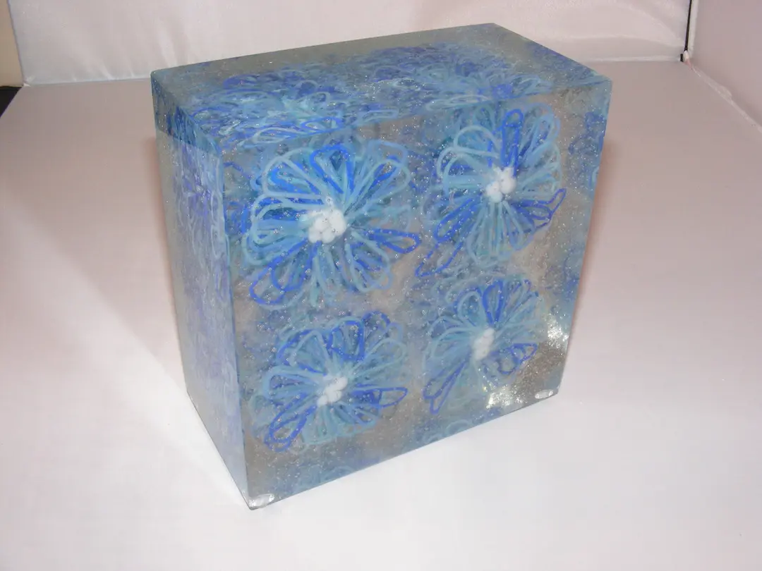 A large, clear glass block with four blue fused glass daisies suspended inside.