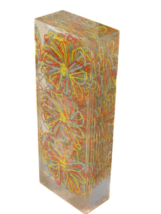 A tall, rectangular glass sculpture with many fused yellow and orange daisy patterns suspended within.