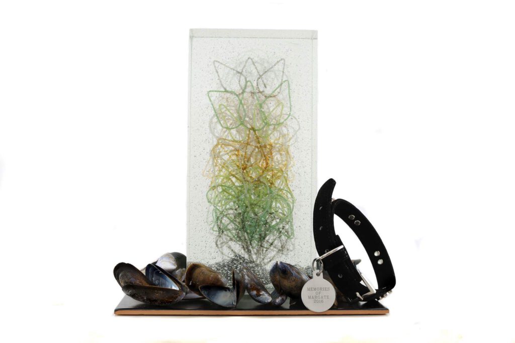 A piece of artwork titled "Memories of Margate" is displayed against a white background. The artwork consists of a clear rectangular block, containing an intricate, abstract design of swirling lines in shades of green and yellow. In front of the block, at its base, several mussel shells rest on a dark, reflective surface that resembles a plinth. Among the shells, a black collar with a circular tag is placed. The tag appears saying Memories of Margate 2016.