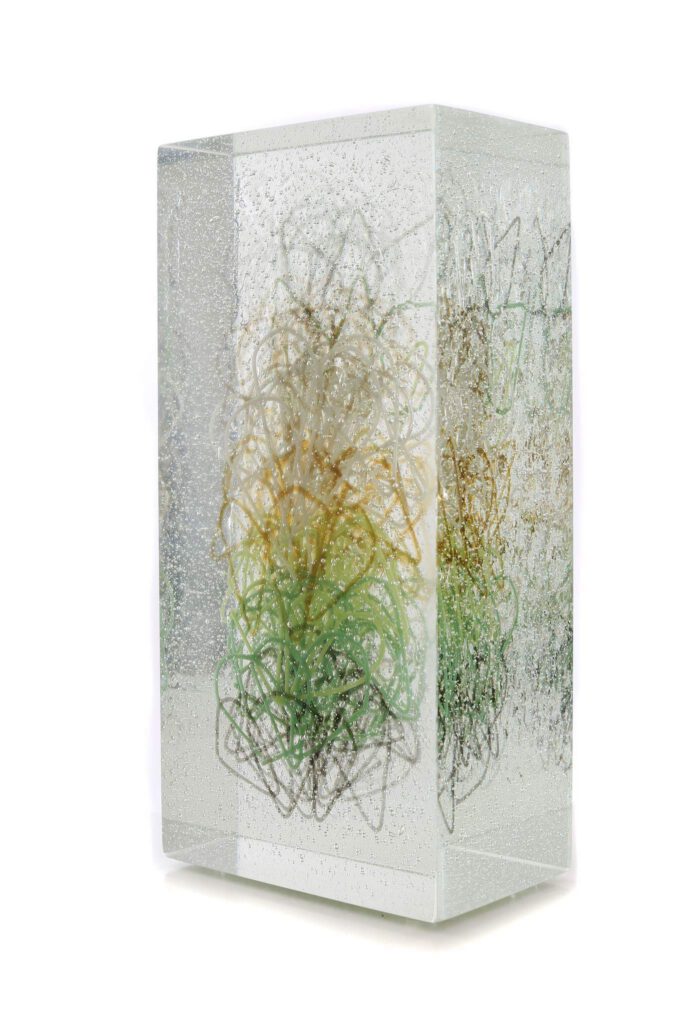 A clear, rectangular glass block, part of the "Memories of Margate" artwork, is shown from a slight angle against a white background. The block is filled with countless tiny air bubbles. Inside the glass, an intricate, abstract design of intertwined, translucent strands.