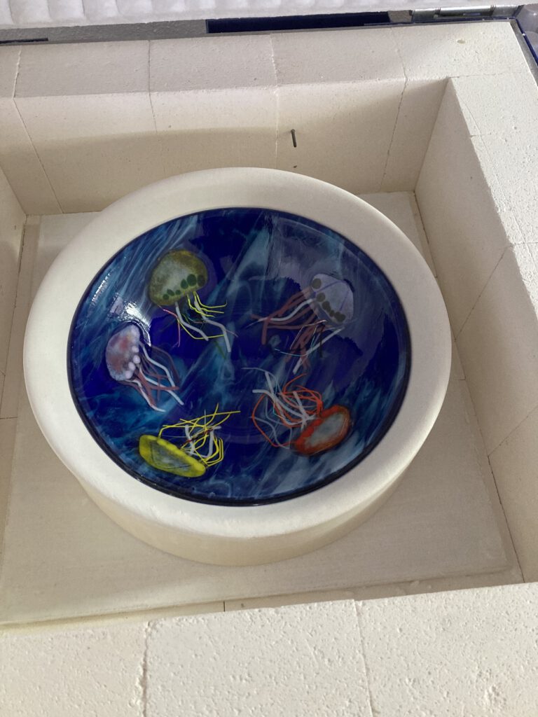 A swirling blue and green abstract glass disc displayed inside a white-rimmed dish.