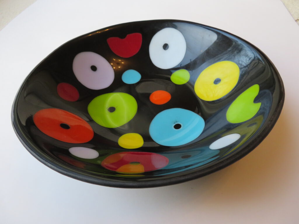 A curved, black glass disc bowl showcasing its playful, multi-coloured circular patterns from a side angle.