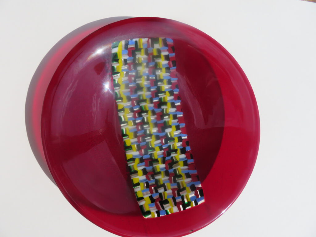 A colourful mosaic panel made of yellow, black, and white glass pieces placed inside a red glass bowl.