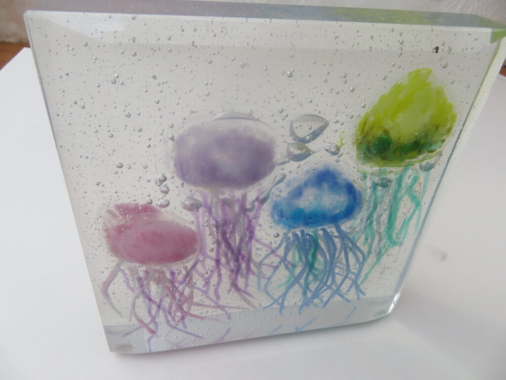 A clear glass box containing three colourful fused glass jellyfish in purple, red, and green.