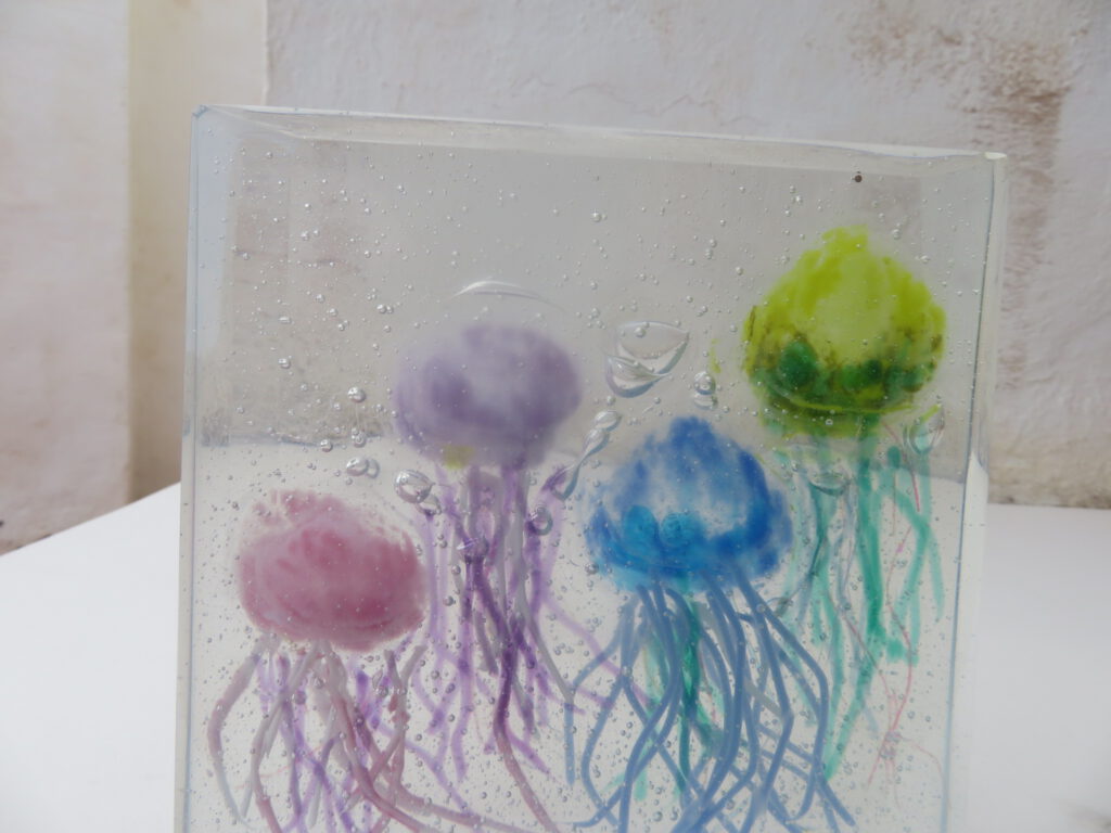 A clear glass box containing three colourful fused glass jellyfish in purple, red, and green.