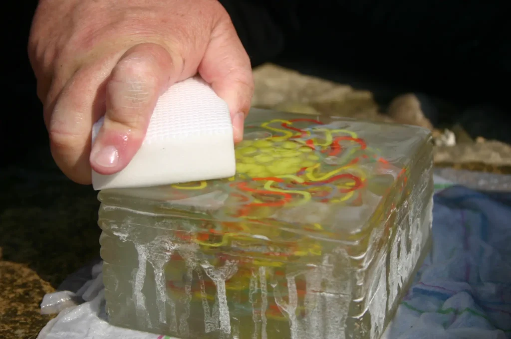 A hand polishing the surface of a large, clear glass block containing a colourful, swirling pattern.