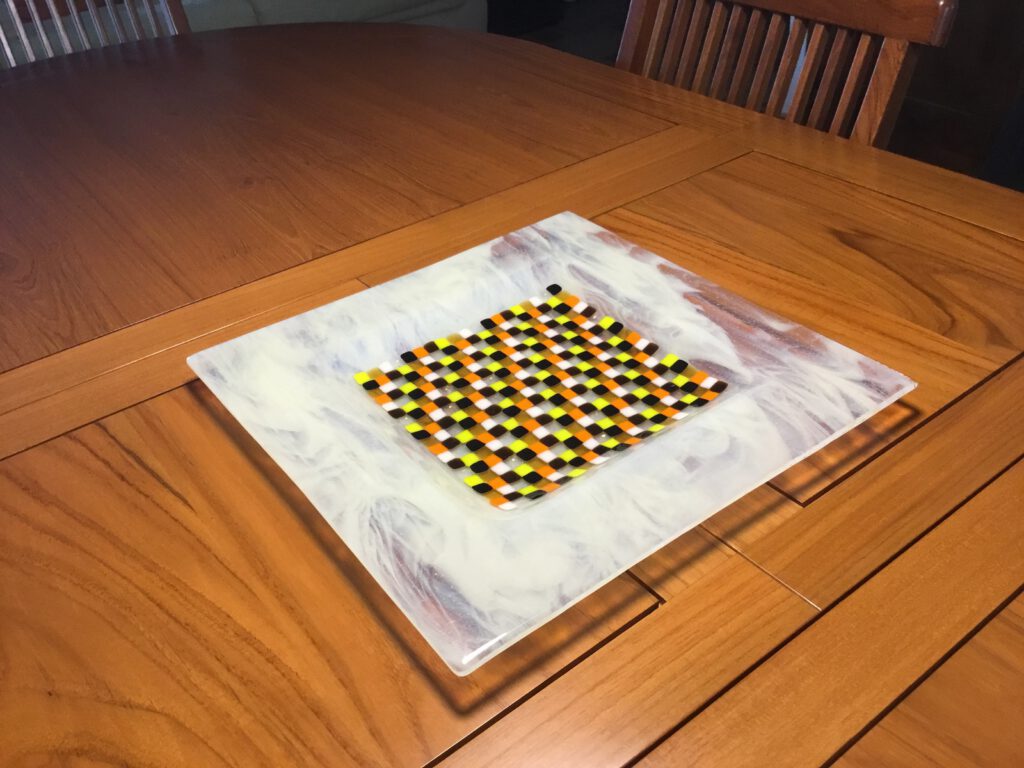 A square glass trivet featuring a central, colourful checkered mosaic pattern on a white marbled glass base.