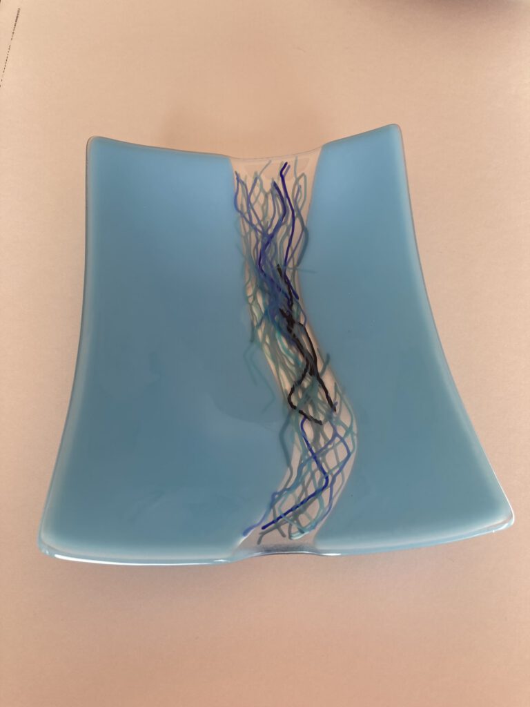 A curved, light blue glass dish with a winding, dark thread-like design resembling a river.