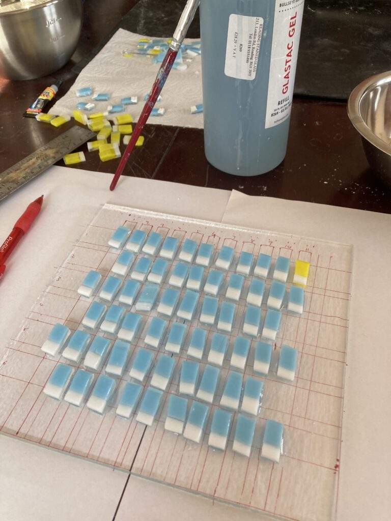 An artist's workspace showing blue and white glass squares being arranged on a clear glass sheet.