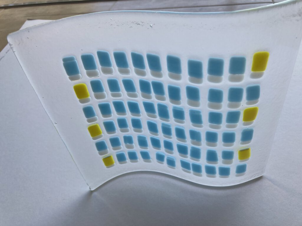 A glass wave sculpture featuring a pattern of fused light blue and white squares with a yellow stripe.