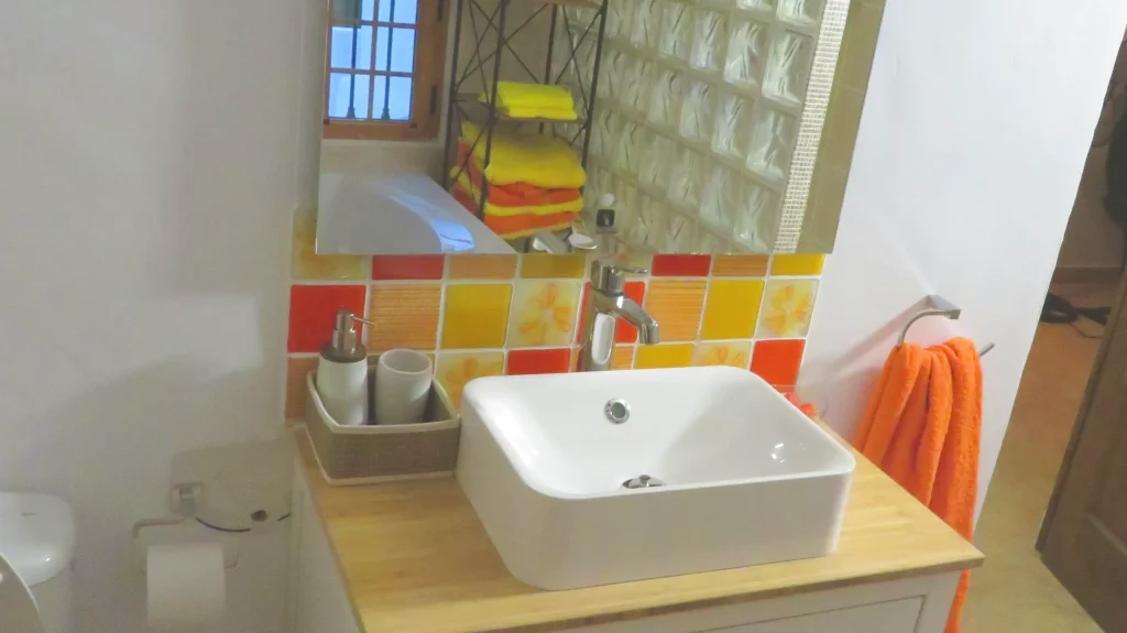 A bathroom vanity area featuring a white square sink on a light wooden countertop. Behind the sink, serving as a splashback, is a vibrant tiled wall of fused glass in various shades of yellow, orange, and red. The tiles have both solid colors and subtle striped or floral patterns. A mirror hangs above the splashback, reflecting a window and stacked orange and yellow towels.