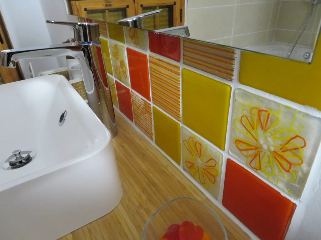 A close-up view of a vibrant fused glass splashback in a bathroom, situated behind a modern white sink and chrome faucet on a light wooden countertop. The splashback is composed of square glass tiles in shades of orange, yellow, and red. Some tiles are solid colored, while others feature delicate etched or fused striped patterns, and a few have subtle floral designs in yellow and orange.