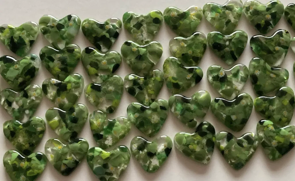 A close-up of many heart-shaped fused glass pieces, predominantly in shades of green with small white and yellow flecks, resembling meadows or 'forget-me-not' flowers. They are tightly grouped together, filling the frame