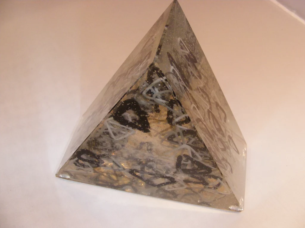 A transparent glass pyramid sculpture, with an opaque base and four triangular faces. Inside the glass are embedded swirling black and white lines, creating abstract patterns, along with many small air bubbles trapped within the glass.