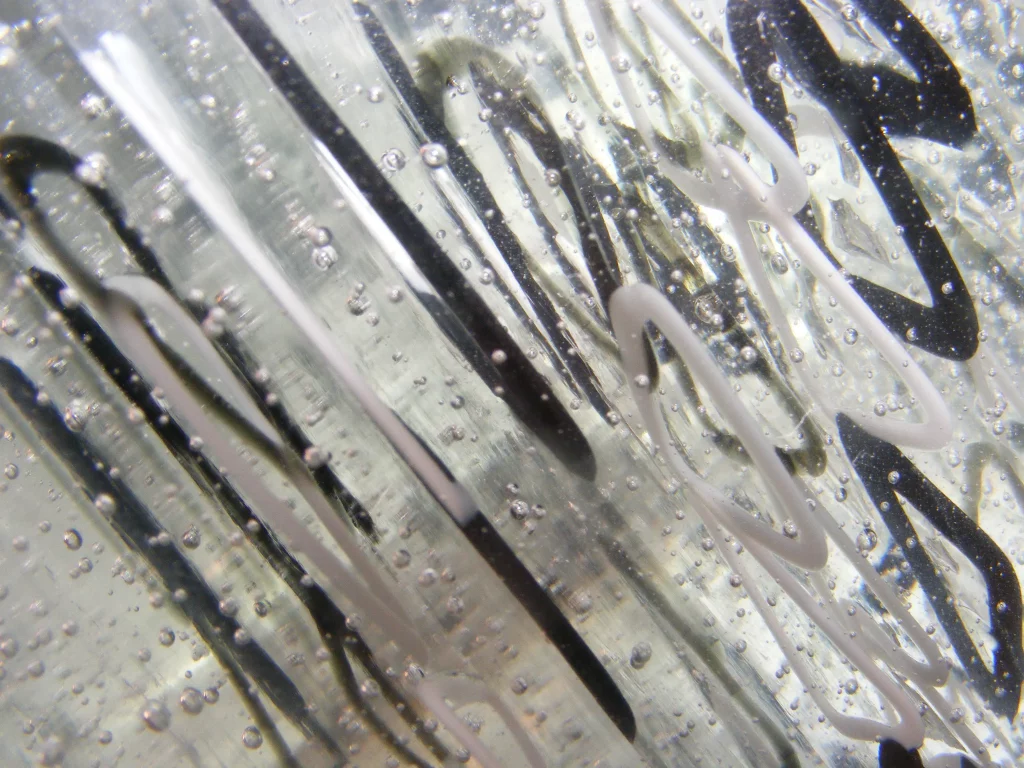 A close-up, abstract view through a piece of clear glass, revealing internal swirling patterns of black and white lines. Many small, round air bubbles are trapped within the glass, creating a textured, almost liquid appearance.