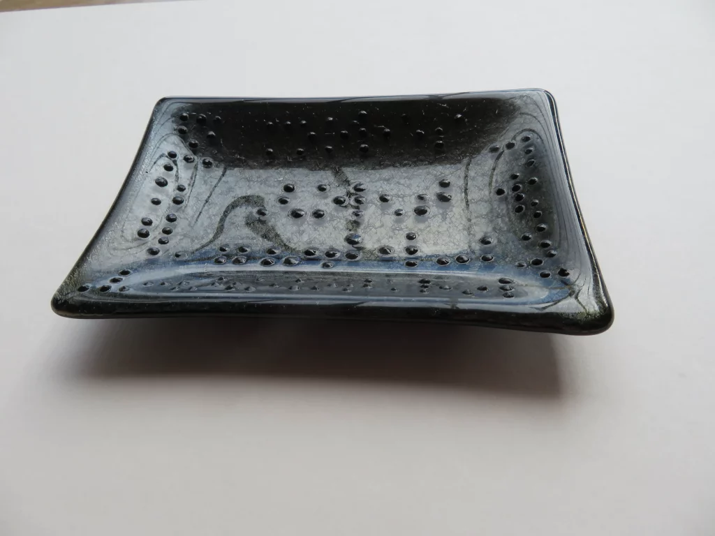 A dark, possibly black or very dark blue, fused glass dish with a textured surface. The dish is rectangular with subtly curved sides, and its surface is covered with many small, raised dots and organic, wavy lines
