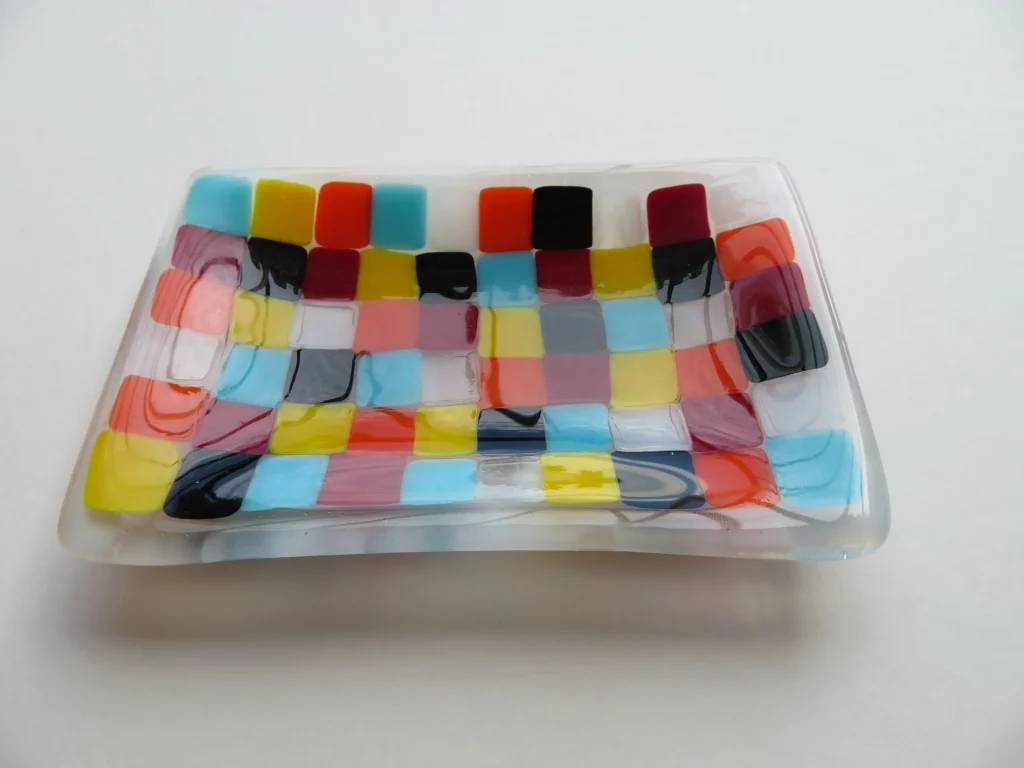 A clear fused glass dish with gently curved sides, adorned with a colorful mosaic pattern of small square and rectangular pieces. The squares are in various bright colors including red, orange, yellow, blue, black, and white, forming a checkered appearance.