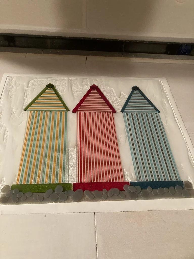 Three flat, rectangular fused glass art panels, each depicting a striped beach hut. From left to right, the huts are green and yellow, red and white, and blue and brown. Each hut has a solid-colored roof and a base of rounded grey glass pieces suggesting pebbles. The panels are on a white surface, likely inside a kiln before firing.