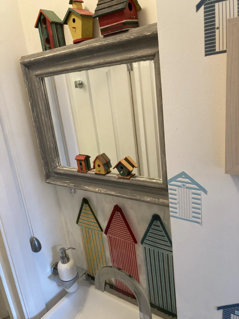 A room interior showing a sink area with a large, framed mirror above it. On the wall above the sink are three large, striped fused glass beach hut panels: green and yellow, red and white, and blue and brown. Reflected in the mirror are three small, colorful wooden birdhouses. A small white pump bottle is visible on the sink to the left.