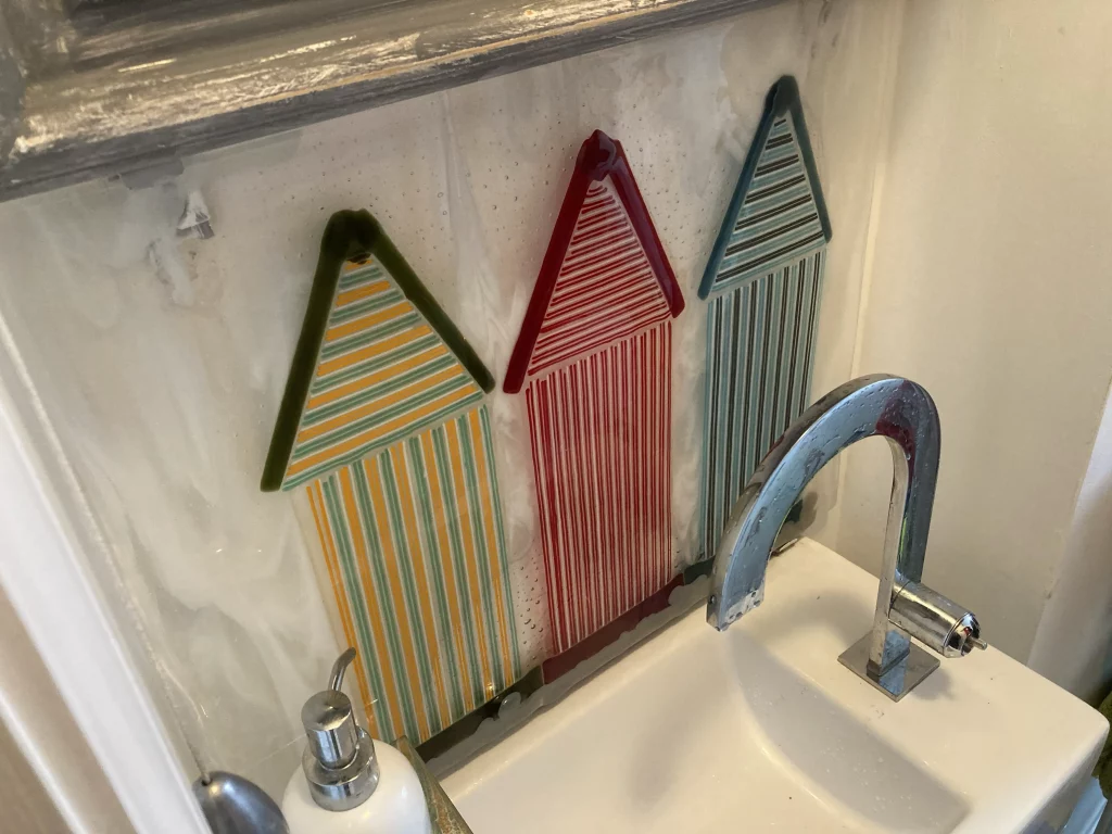 A close-up view of three fused glass beach hut art panels mounted on a wall above a white sink with a chrome faucet. The huts are striped in green and yellow, red and white, and blue and brown. The glass reflects light from the surrounding room.