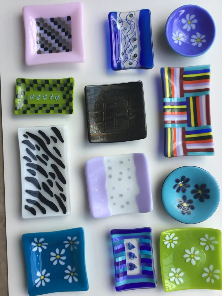 Twelve small, various shaped fused glass dishes arranged in three rows on a white surface. Designs include checkered patterns, abstract swirls, vertical stripes, and daisy flowers in a range of colors like purple, blue, green, black, and white.
