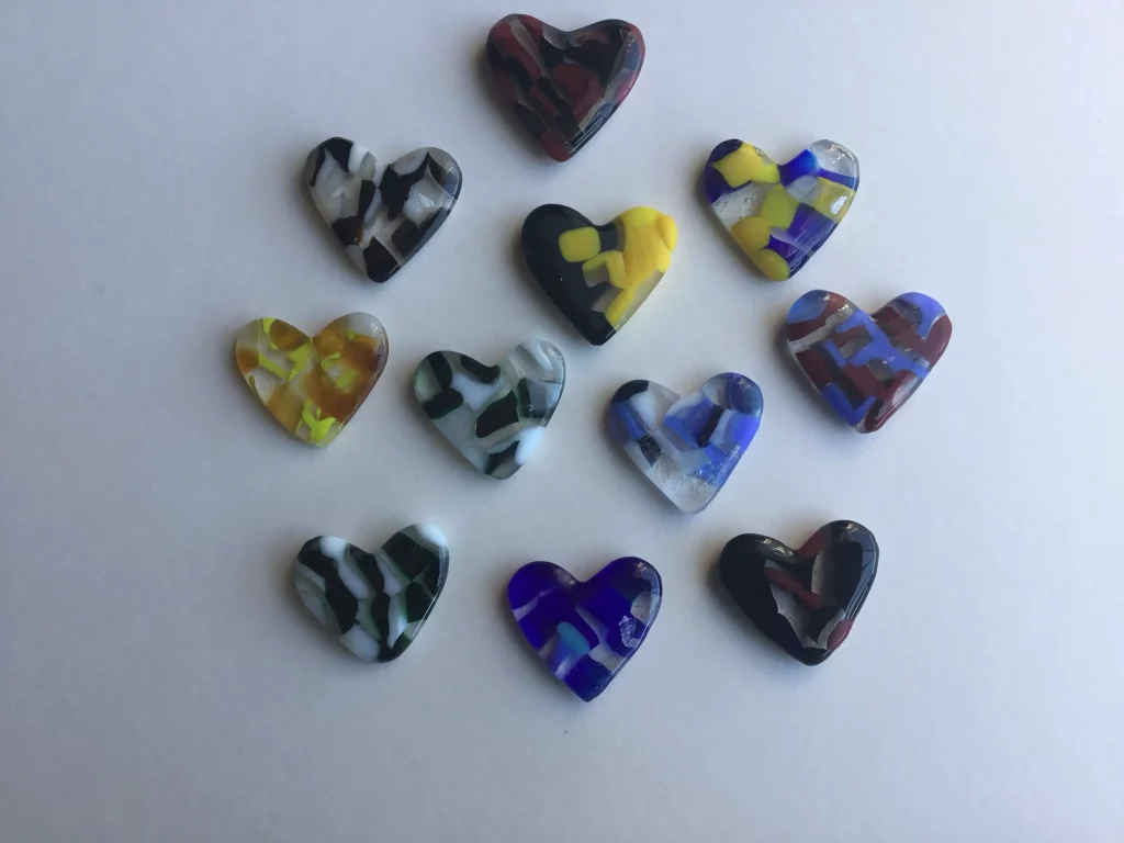Eleven various fused glass hearts arranged on a white surface, forming a loose triangular shape with one heart at the top. Each heart displays different multi-colored abstract designs.
