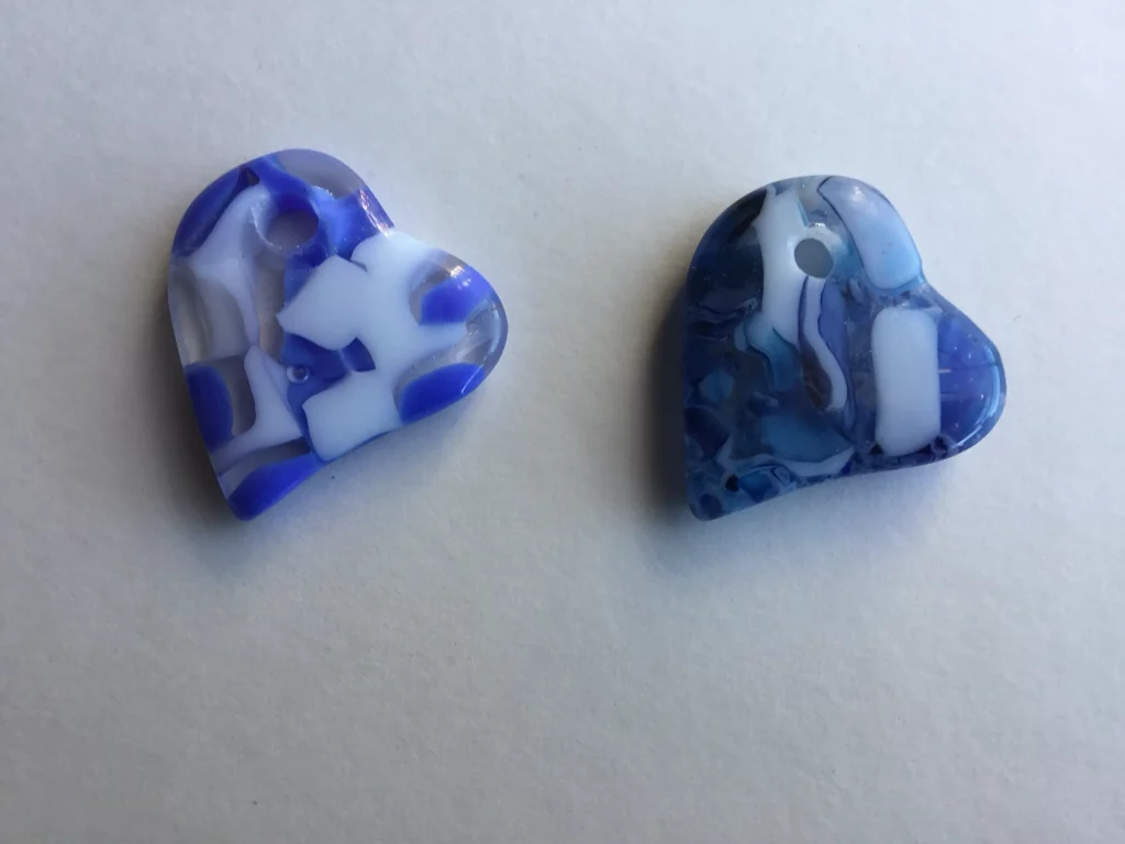 Two blue and white fused glass hearts on a white surface. The heart on the left has clear and bright blue glass with distinct white shapes. The heart on the right is a darker blue with more blended white and light blue patterns.
