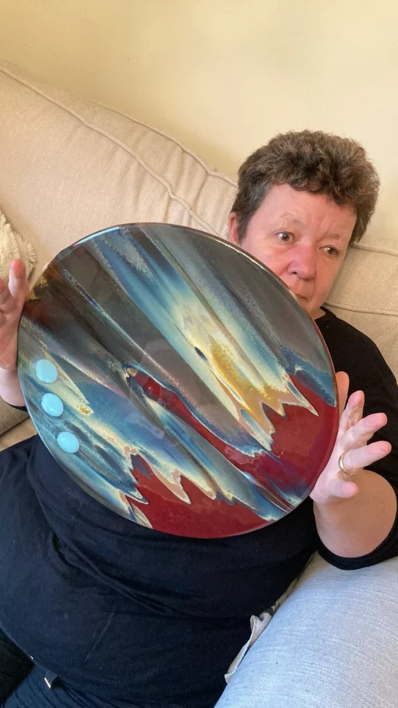 Julie Coakley, holding a large, round fused glass plate titled 'Petrified Wood' in front of her. The plate features swirling blues, browns, and reds with a textured, wood-like appearance, and three light blue circular inclusions on the left side.