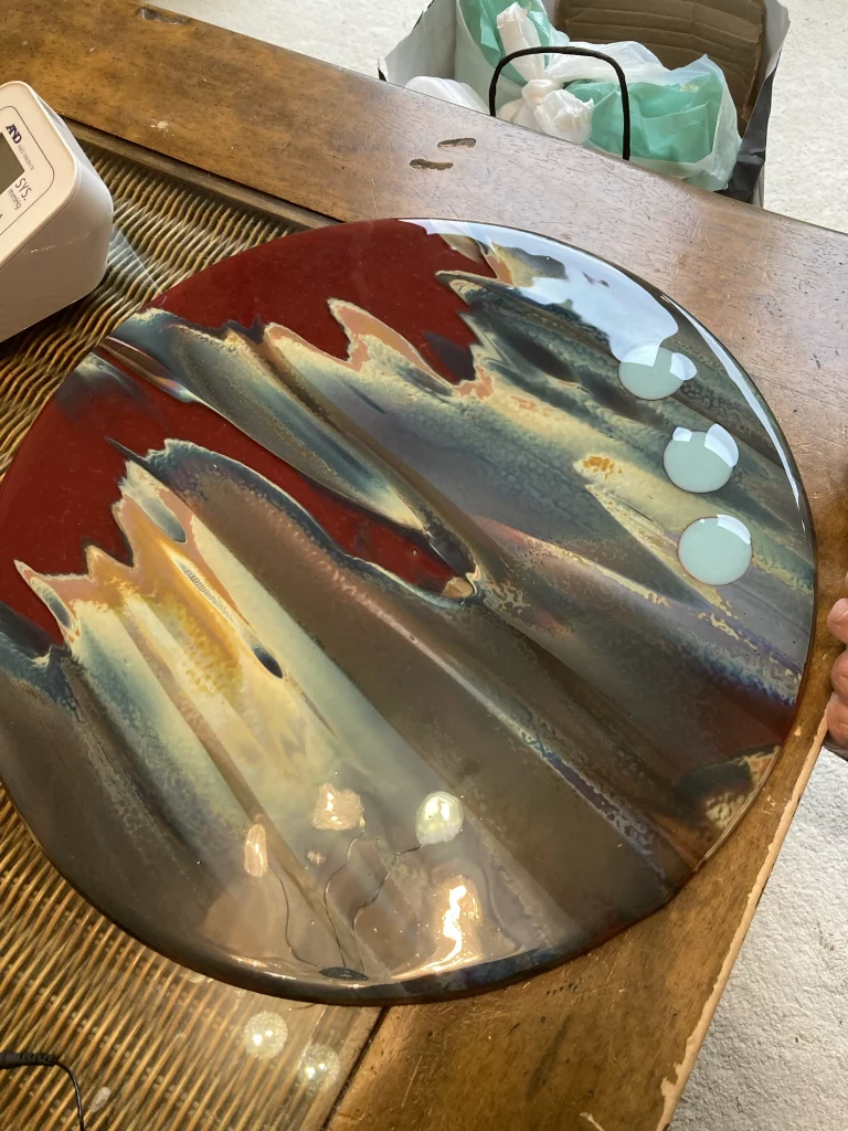 A large, round fused glass plate titled 'Petrified Wood' resting on a textured brown wooden table or surface. The plate features a prominent design of rich reds, blues, greens, and browns in swirling, textured patterns, characteristic of petrified wood. Three light blue circular inclusions are visible on the right side.
