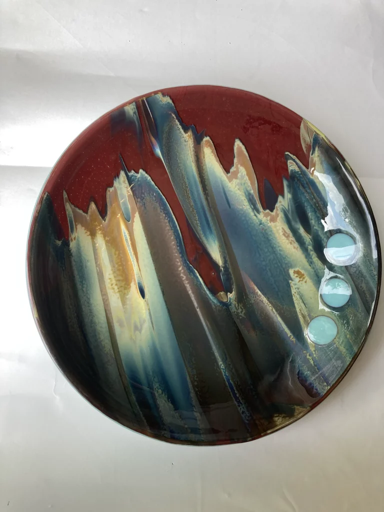 A large, round fused glass plate titled 'Petrified Wood,' lying flat on a white surface. The plate features a dynamic abstract design with rich, dark red sections contrasted by shimmering blues, greens, and golden browns that flow in wavy, textured bands, reminiscent of wood grain. Three small, light blue circular inclusions are visible on the right side.