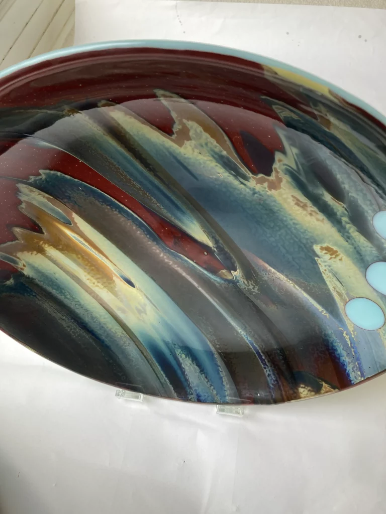 An angled view of a large, round fused glass plate titled 'Petrified Wood,' displayed upright on a clear stand against a white background. The plate's abstract design showcases a vibrant interplay of deep reds, blues, greens, and shimmering goldens, with distinct textured lines resembling natural wood grain. Three light blue circular inclusions are visible on the right.