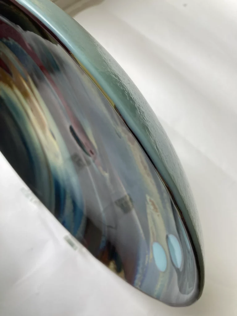 A close-up, angled view of the edge of a large, round fused glass plate titled 'Petrified Wood.' The surface shows a blend of shimmering blues, greens, and browns flowing into a dark red section, with the smooth, slightly raised edge of the plate visible in a muted teal color. Two light blue circular inclusions are visible on the right.