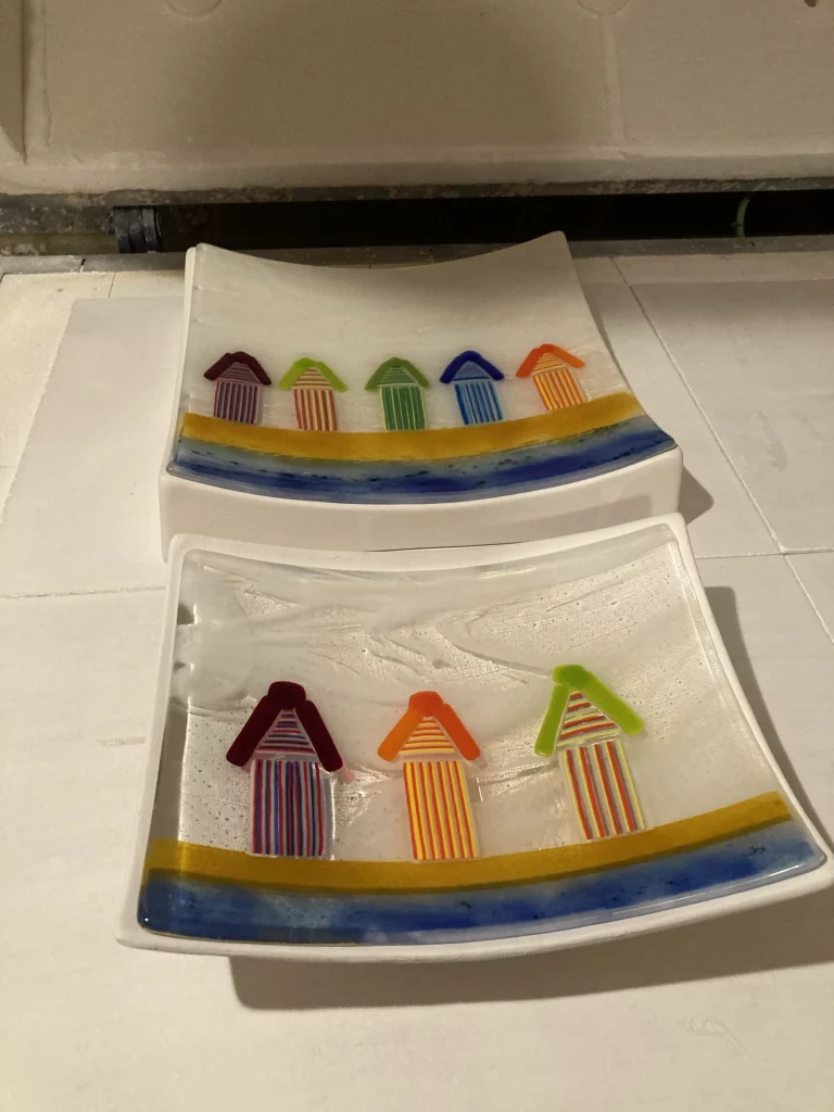 Two rectangular fused glass dishes, each featuring a wavy yellow stripe above a blue stripe, creating a beach scene. On the yellow stripe of the top dish are five small, colorful beach huts: a purple, lime green, dark green, blue, and orange hut. The bottom dish has three similar huts: a red, orange, and lime green hut.