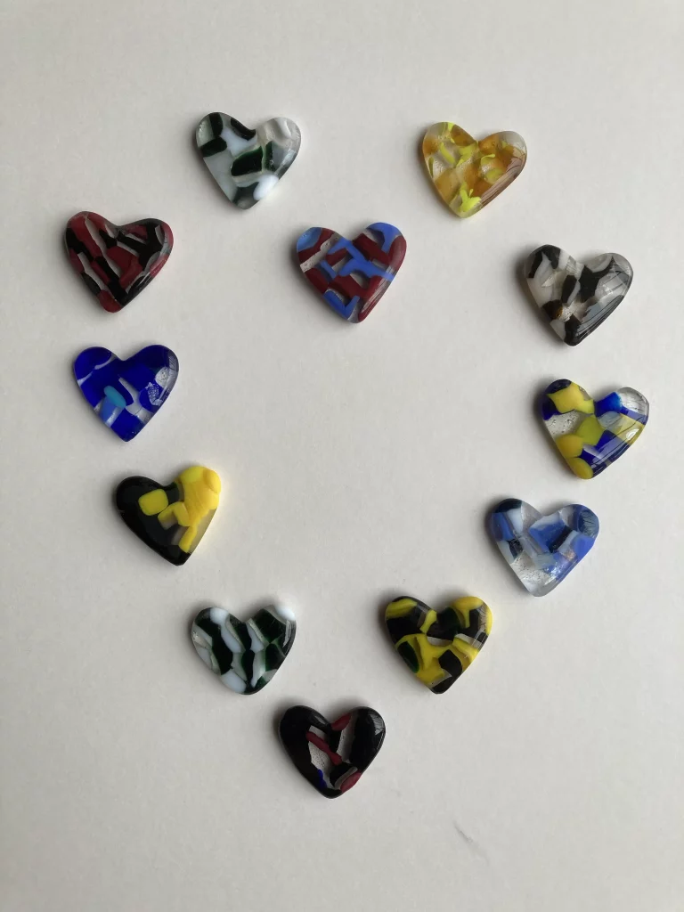 Eleven diverse, heart-shaped pieces of fused glass, arranged in a semicircle on a white background. Each heart features unique vibrant color combinations and abstract patterns.