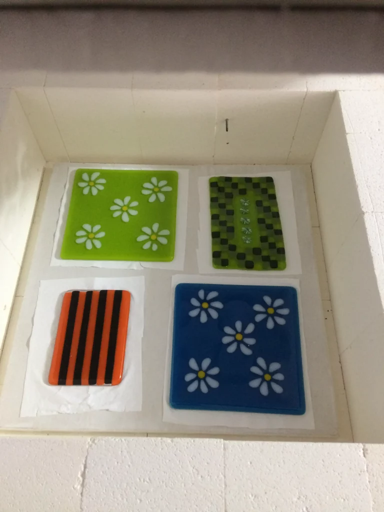 Four rectangular fused glass panels or flat dishes, arranged on white paper inside a kiln, ready for firing. The top left is lime green with white daisies. The top right is green and black checkered. The bottom left has bold orange and black vertical stripes. The bottom right is blue with white daisies.