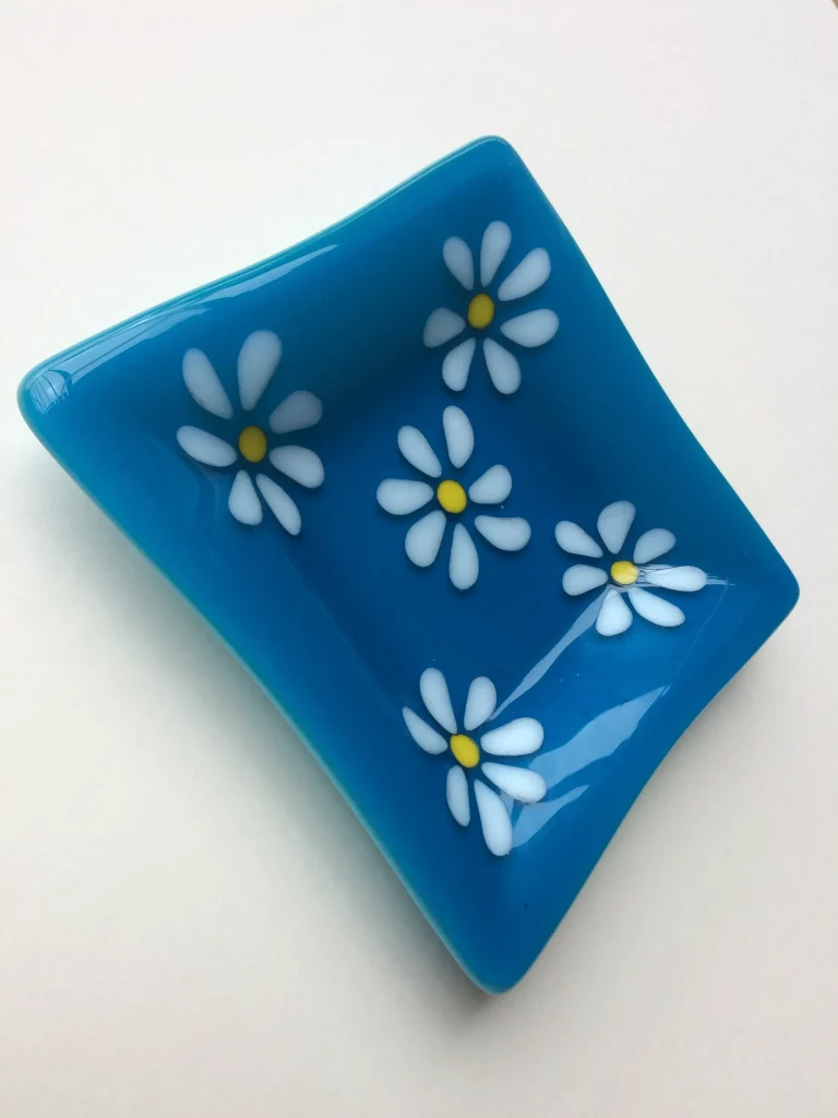 A small, deep blue fused glass dish with gently curved sides. Five white daisy flowers with yellow centers are fused onto its surface, arranged to spread across the dish.