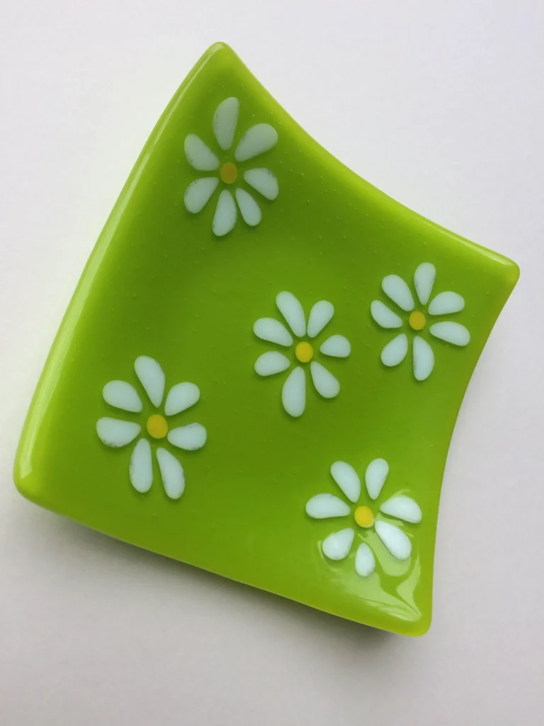 A small, vibrant lime green fused glass dish with gently curved sides. Five white daisy flowers with yellow centers are fused onto its surface, arranged to spread across the dish