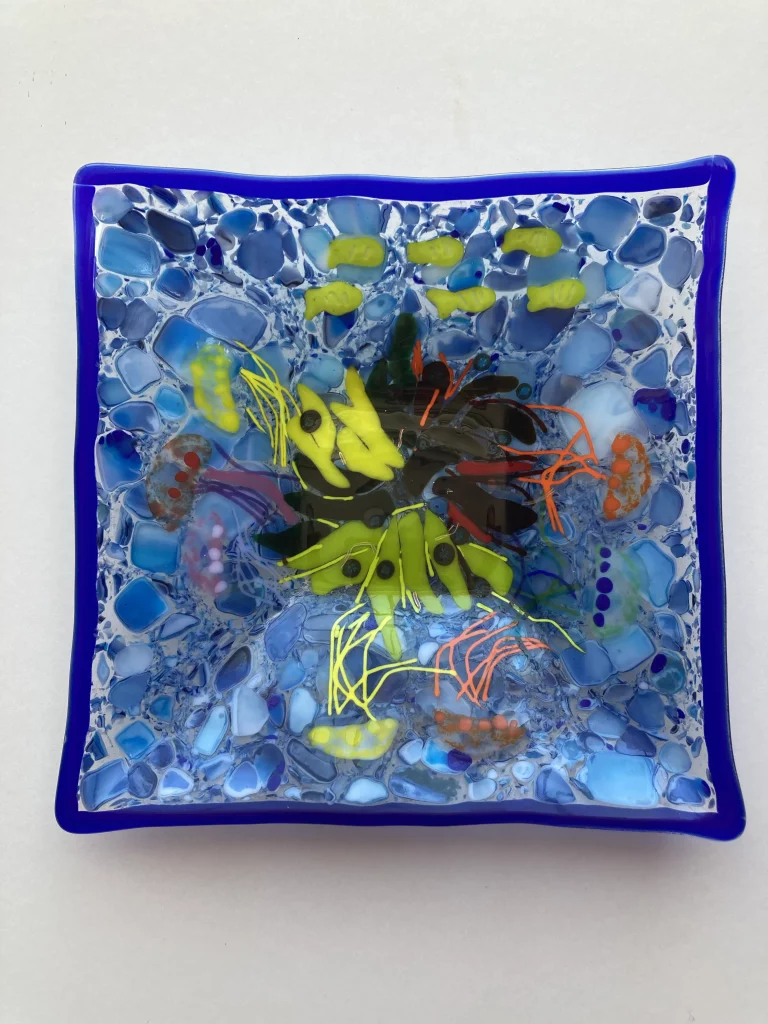 A square, deep blue-edged fused glass dish depicting an abstract underwater scene. The center features a swirl of green and black elements with bright yellow and orange lines, surrounded by various shades of blue glass pieces resembling water and bubbles.