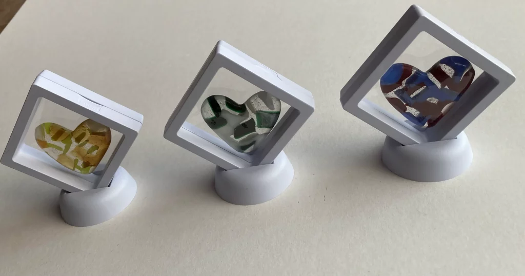 Three colorful fused glass hearts, each displayed inside a clear, square plastic frame with a white base that tilts the heart upwards. From left to right: a yellow and green heart, a green and white heart, and a blue, brown, and white heart.