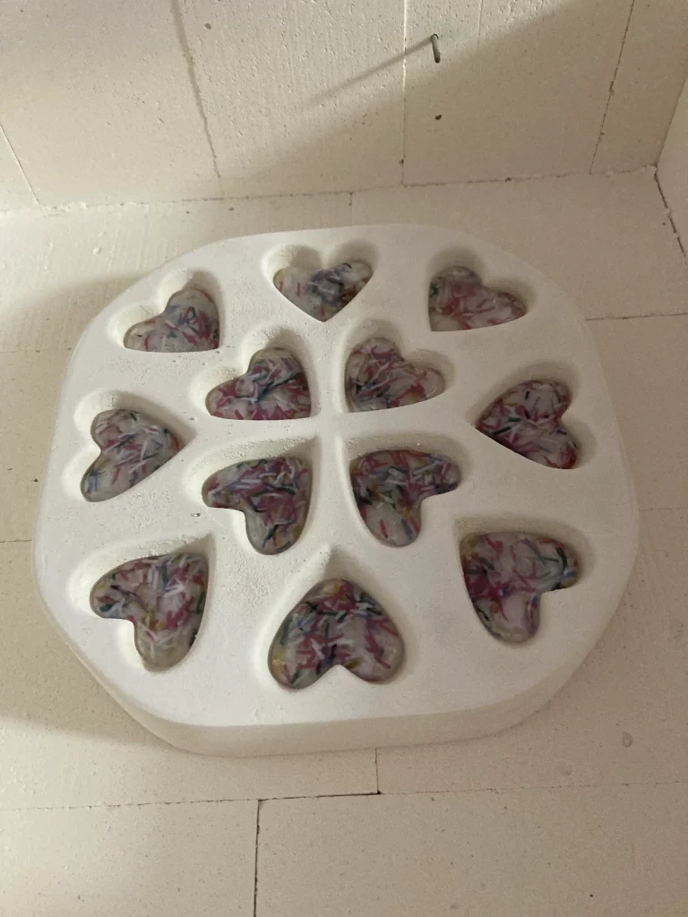Ten colorful, heart-shaped pieces of fused glass, each with unique swirling patterns, resting in the indentations of a white ceramic mold inside a kiln, ready for firing.